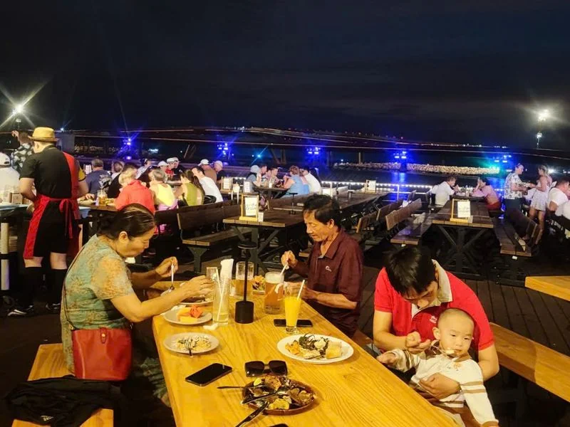 Gia đình ba thế hệ gồm bà, ông và bố bế em bé ngồi ăn buffet tại bàn gỗ dài nhà hàng Sun Bavaria